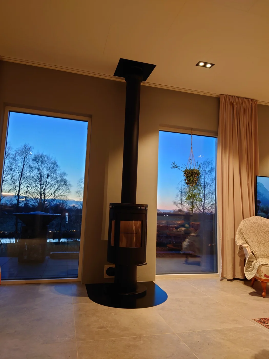 Svart kamin framför stora glaspartier i skymningsljus